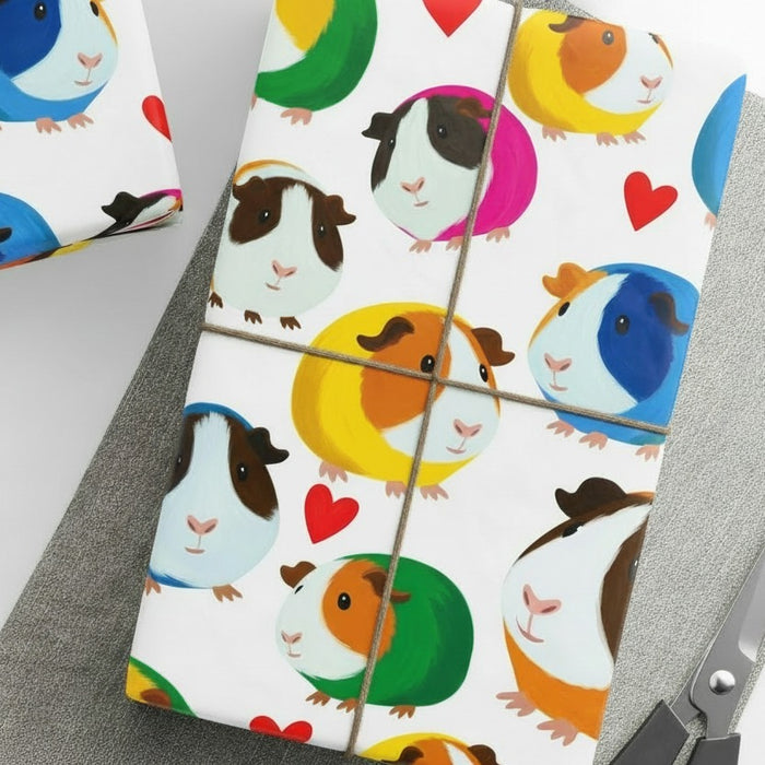 A gift box wrapped in white wrapping paper printed with a colorful cartoon guinea pig pattern and small red hearts, photographed on a gray surface with scissors partially visible at the corner 